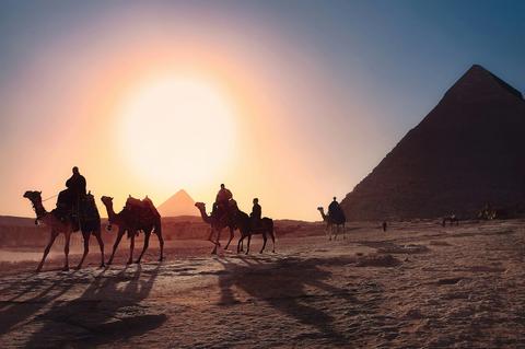 Kamelreiter ziehen bei Sonnenuntergang vor den Pyramiden durch den Sand